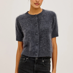 A woman modeling a short-sleeve fuzzy knit cardigan in charcoal gray with dark buttons, paired with black jeans, against a plain light gray studio background.