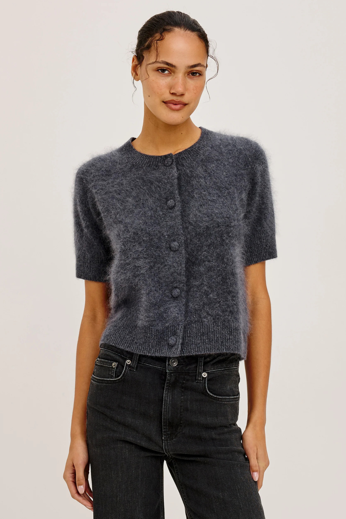 A woman modeling a short-sleeve fuzzy knit cardigan in charcoal gray with dark buttons, paired with black jeans, against a plain light gray studio background.