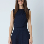 A woman with long brown hair and glasses wearing a sleeveless dark navy blue dress, standing against a light gray studio background.