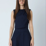 A woman with long brown hair and glasses wearing a sleeveless dark navy blue dress, standing against a light gray studio background.