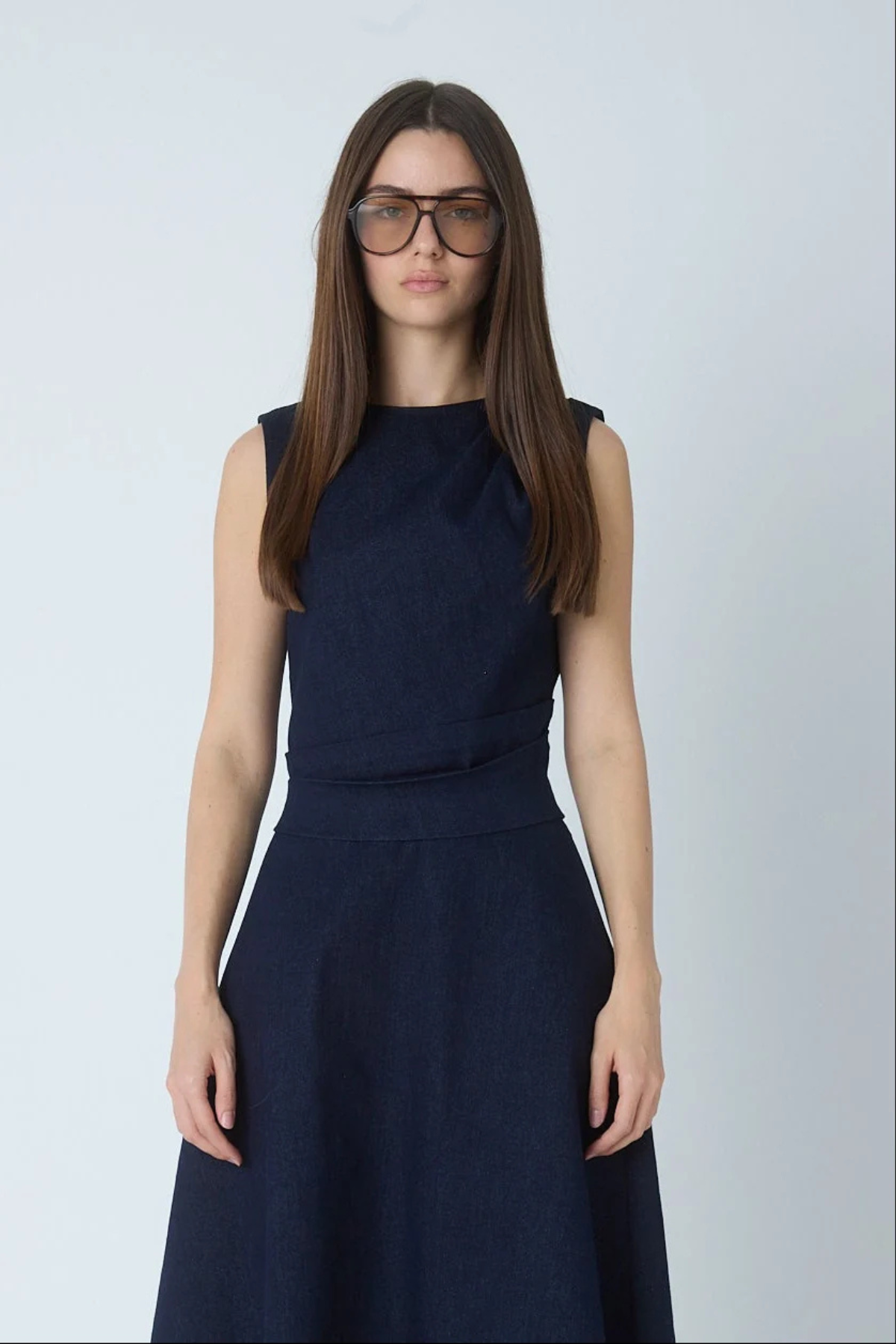 A woman with long brown hair and glasses wearing a sleeveless dark navy blue dress, standing against a light gray studio background.