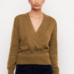 A woman modeling a brown knit wrap sweater with a V-neck and long sleeves, paired with dark pants, against a plain light background.