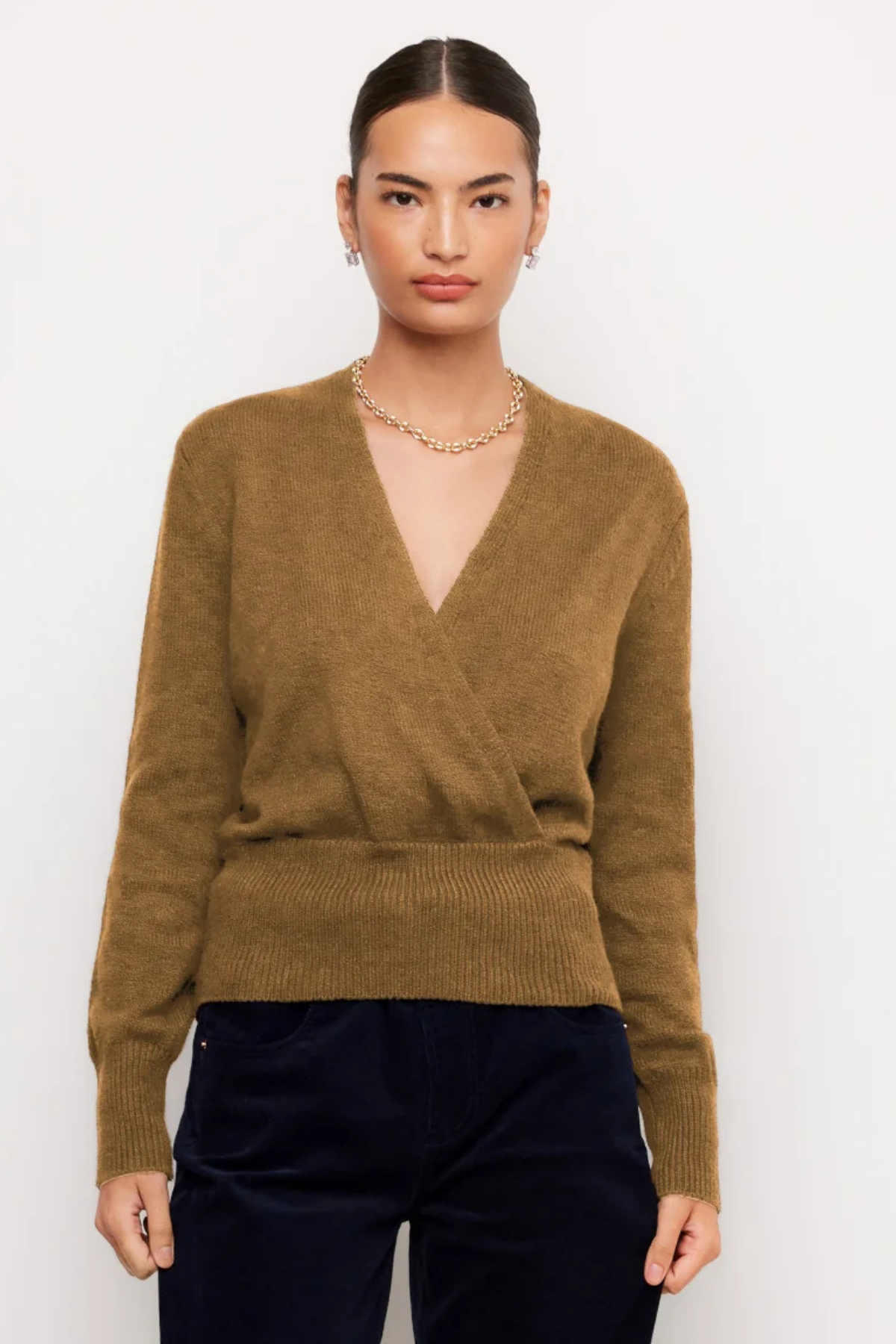 A woman modeling a brown knit wrap sweater with a V-neck and long sleeves, paired with dark pants, against a plain light background.