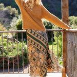 A woman stands on a wooden deck wearing a fuzzy mustard-brown sweater and a long patterned skirt with green, gold, and brown tones. She is posing outdoors with a scenic garden background.