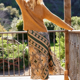 A woman stands on a wooden deck wearing a fuzzy mustard-brown sweater and a long patterned skirt with green, gold, and brown tones. She is posing outdoors with a scenic garden background.