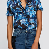 A woman wearing a short-sleeve button-front blouse with a blue and white floral print on a black background, paired with high-waisted blue jeans featuring black lace side panels, against a light gray studio background.