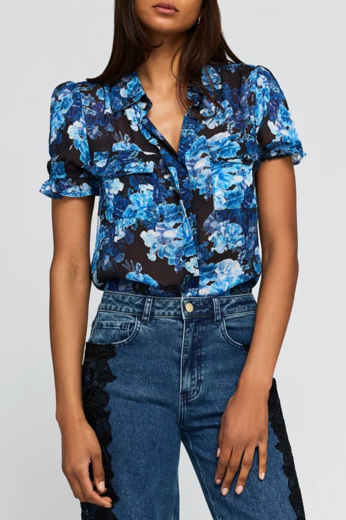 A woman wearing a short-sleeve button-front blouse with a blue and white floral print on a black background, paired with high-waisted blue jeans featuring black lace side panels, against a light gray studio background.
