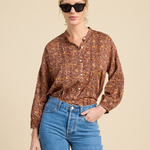 A woman modeling a long-sleeve button-up shirt with a dense rust-brown, orange, and yellow floral pattern, paired with blue jeans and black sunglasses, against a solid beige background.