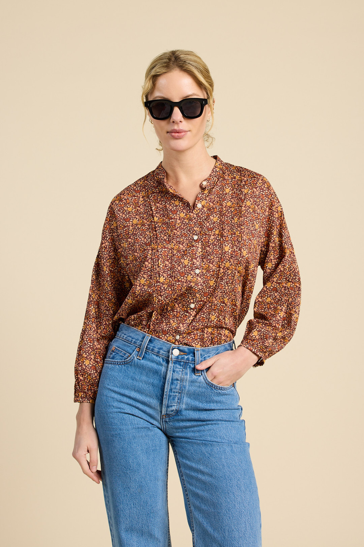 A woman modeling a long-sleeve button-up shirt with a dense rust-brown, orange, and yellow floral pattern, paired with blue jeans and black sunglasses, against a solid beige background.