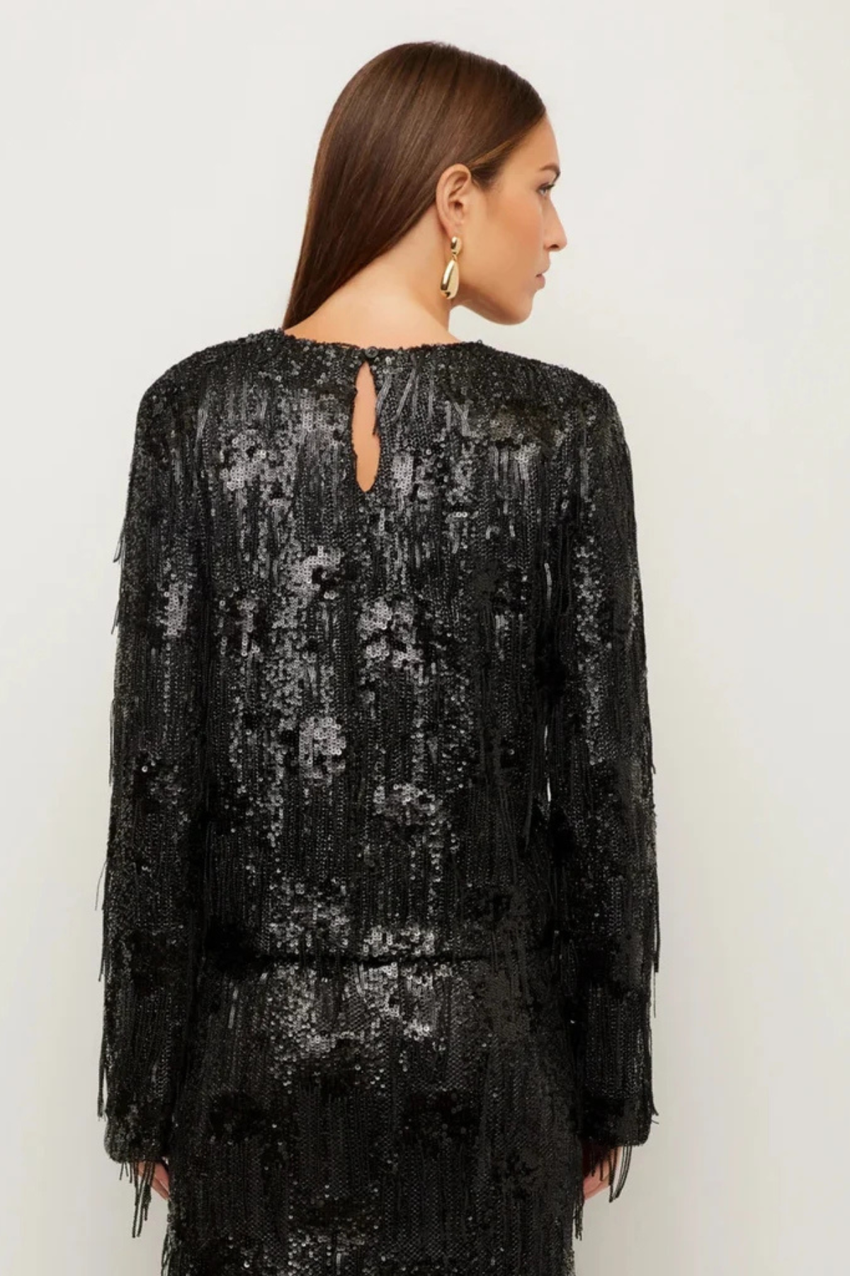 A woman shown from the back wearing a black long-sleeve sequined dress with fringe detailing and a small keyhole cutout at the neckline, against a plain light background.