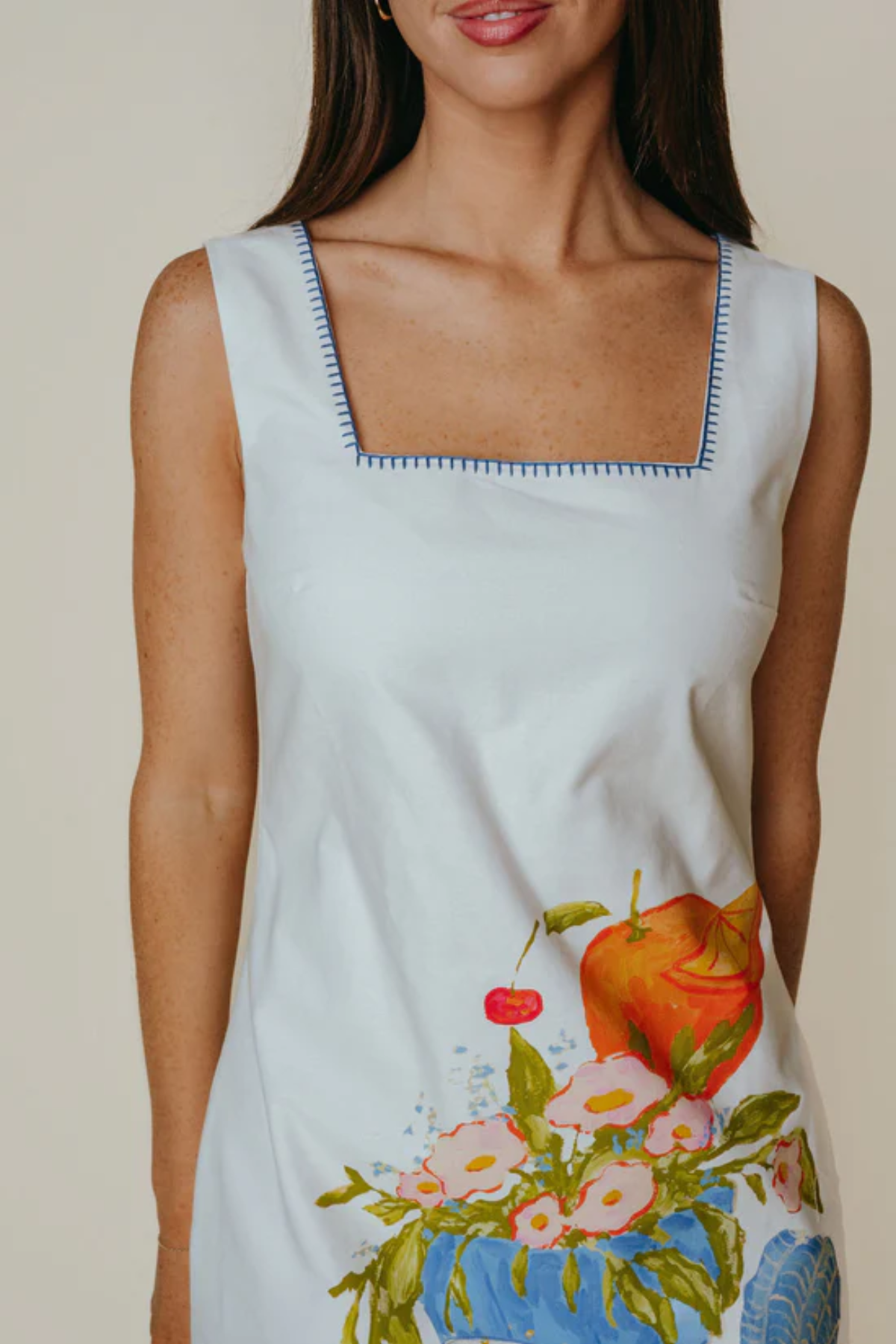 Woman wearing a sleeveless dress with a floral and fruit design on a plain background