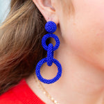 Close-up of a person wearing blue beaded earrings with a red shirt and gold necklace.