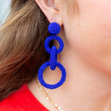 Close-up of a person wearing blue beaded earrings with a red shirt and gold necklace.