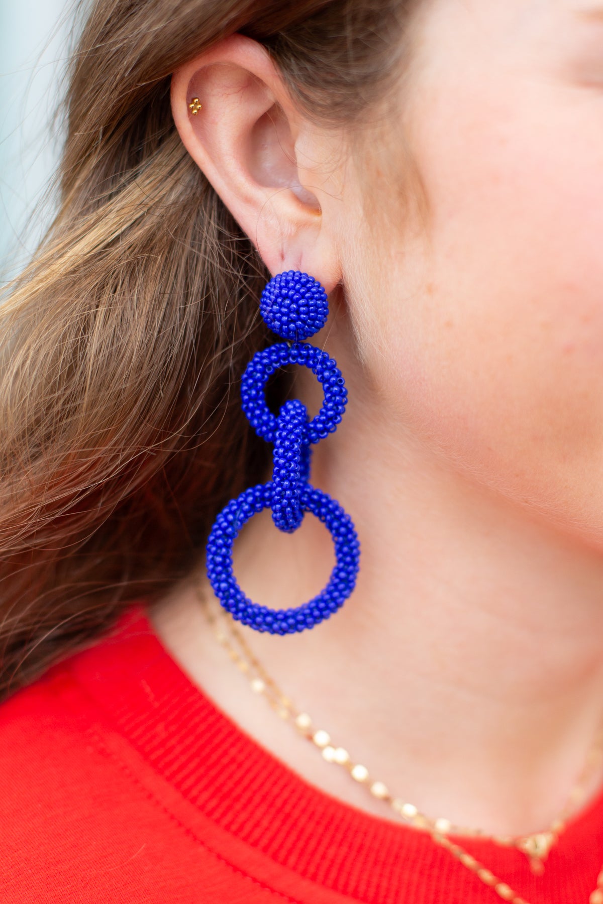 Close-up of a person wearing blue beaded earrings with a red shirt and gold necklace.