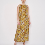 A woman modeling a sleeveless, floor-length maxi dress with a bold yellow, gold, and brown floral medallion pattern against a plain white studio background.