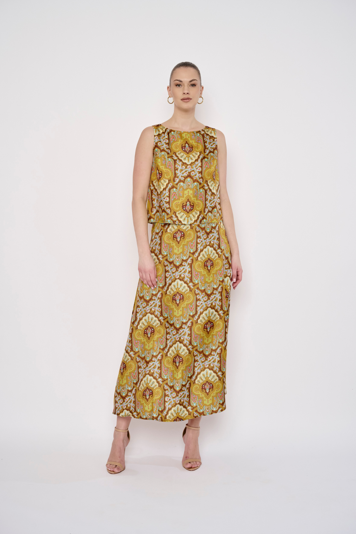 A woman modeling a sleeveless, floor-length maxi dress with a bold yellow, gold, and brown floral medallion pattern against a plain white studio background.