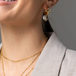 Close-up of a woman wearing a gold hoop earring with a white crescent moon charm, layered with gold necklaces, wearing a light gray blazer.