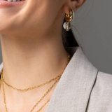 Close-up of a woman wearing a gold hoop earring with a white crescent moon charm, layered with gold necklaces, wearing a light gray blazer.