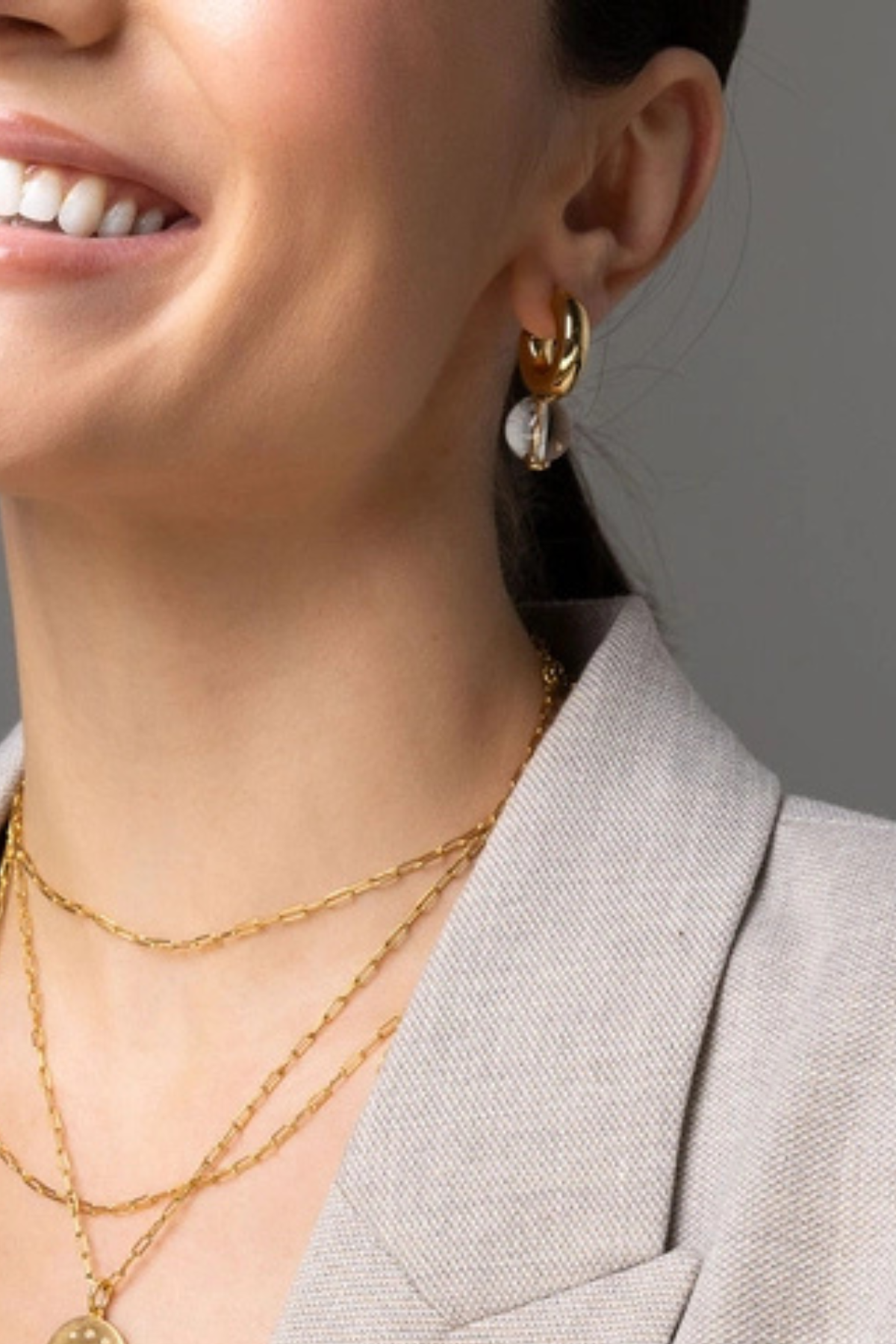Close-up of a woman wearing a gold hoop earring with a white crescent moon charm, layered with gold necklaces, wearing a light gray blazer.