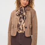 A model wearing a cropped taupe-brown suede jacket, a patterned beige and brown scarf, and dark brown trousers, photographed against a clean light gray studio background.