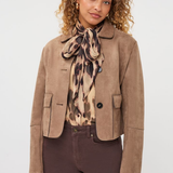 A model wearing a cropped taupe-brown suede jacket, a patterned beige and brown scarf, and dark brown trousers, photographed against a clean light gray studio background.