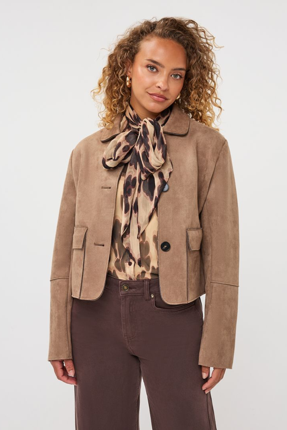 A model wearing a cropped taupe-brown suede jacket, a patterned beige and brown scarf, and dark brown trousers, photographed against a clean light gray studio background.