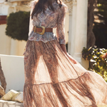 Woman in a long, patterned dress with a belt standing outdoors.