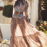 Woman in a long, patterned dress with a belt standing outdoors.