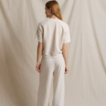 A woman stands with her back to the camera wearing a matching cream-colored short-sleeve top and wide-leg trousers set, posed against a draped cream fabric backdrop.