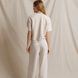 A woman stands with her back to the camera wearing a matching cream-colored short-sleeve top and wide-leg trousers set, posed against a draped cream fabric backdrop.