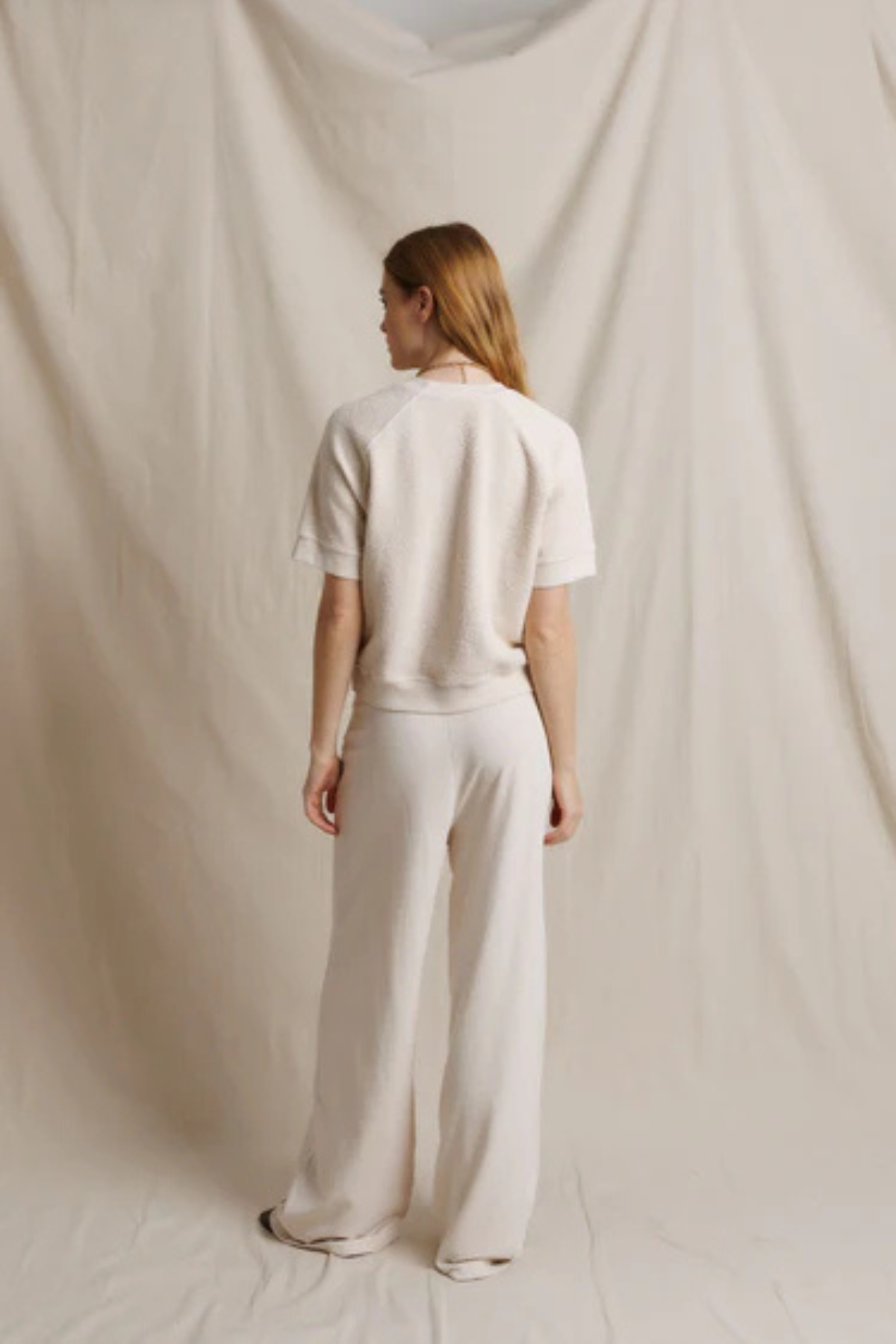 A woman stands with her back to the camera wearing a matching cream-colored short-sleeve top and wide-leg trousers set, posed against a draped cream fabric backdrop.