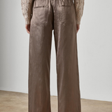 A person shown from the back wearing wide-leg taupe pants and a beige knit sweater, standing against a plain light gray wall and concrete floor.