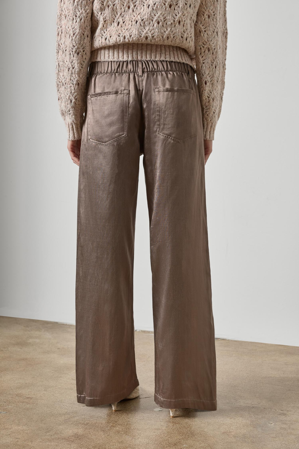 A person shown from the back wearing wide-leg taupe pants and a beige knit sweater, standing against a plain light gray wall and concrete floor.