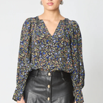 A woman modeling a long-sleeve V-neck blouse with a dense multicolor floral print in blue, yellow, and green on a black base, paired with a black leather-look button-front skirt against a light gray studio background.