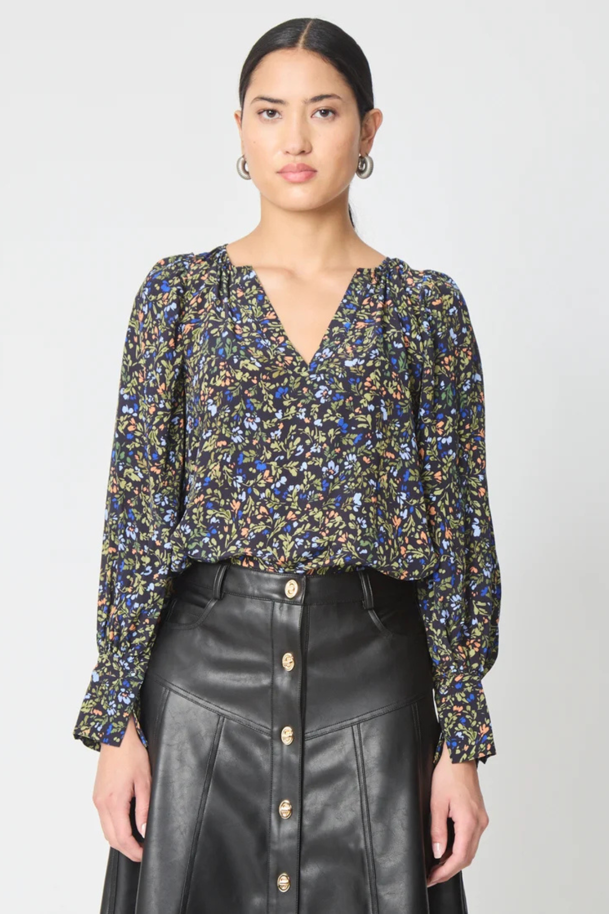 A woman modeling a long-sleeve V-neck blouse with a dense multicolor floral print in blue, yellow, and green on a black base, paired with a black leather-look button-front skirt against a light gray studio background.