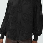 A black long-sleeve button-up blouse with tonal floral embroidery and sheer mesh details, worn by a model with brown pants against a light gray background.
