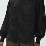 A black long-sleeve button-up blouse with tonal floral embroidery and sheer mesh details, worn by a model with brown pants against a light gray background.