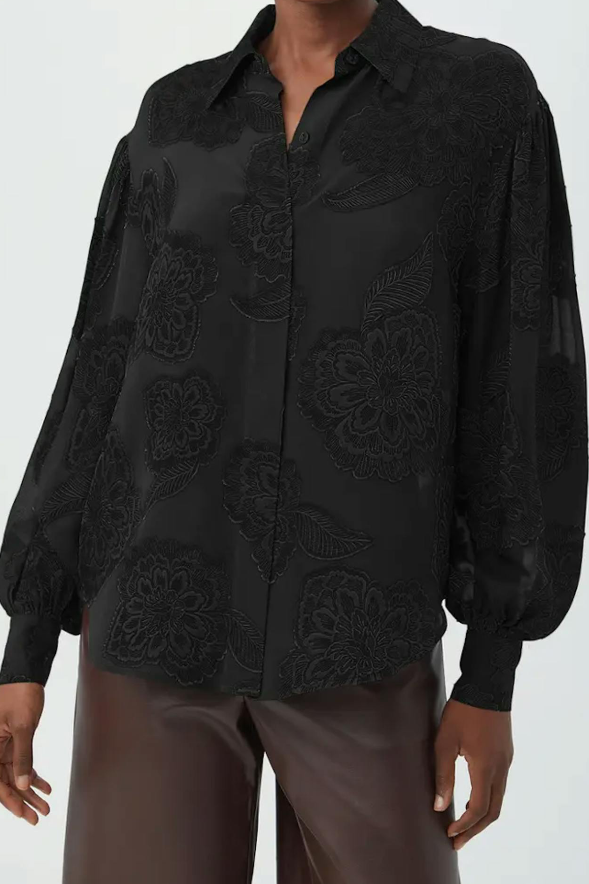 A black long-sleeve button-up blouse with tonal floral embroidery and sheer mesh details, worn by a model with brown pants against a light gray background.