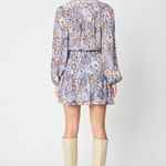 A woman shown from the back wearing a long-sleeve mini dress with a blue, orange, and beige paisley pattern, paired with beige knee-high boots, against a light gray studio background.