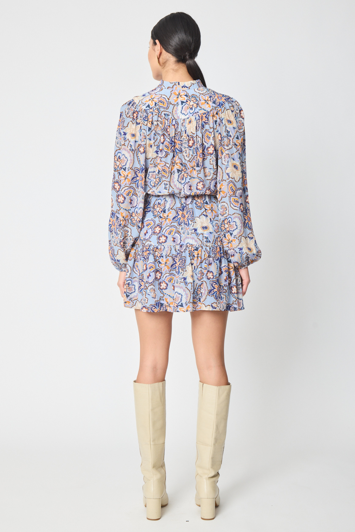 A woman shown from the back wearing a long-sleeve mini dress with a blue, orange, and beige paisley pattern, paired with beige knee-high boots, against a light gray studio background.