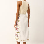 Woman wearing a cream top and white skirt with floral embroidery on a plain background