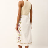 Woman wearing a cream top and white skirt with floral embroidery on a plain background