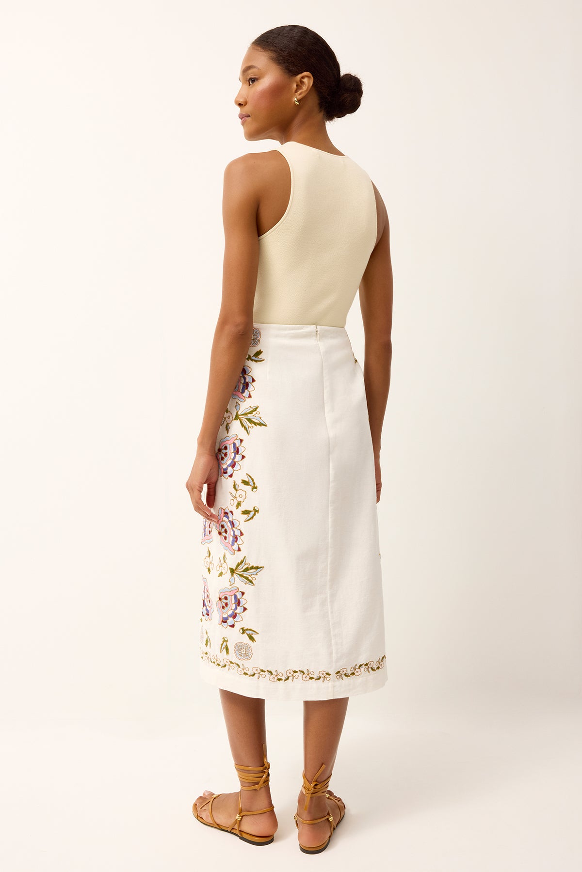 Woman wearing a cream top and white skirt with floral embroidery on a plain background