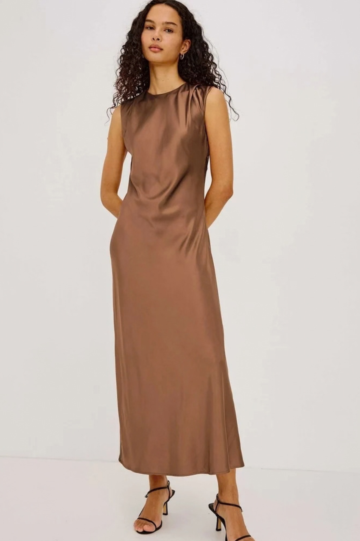 A woman modeling a sleeveless, midi-length brown satin dress with a high neckline and gathered shoulders, paired with black strappy high-heeled sandals against a plain white studio background.