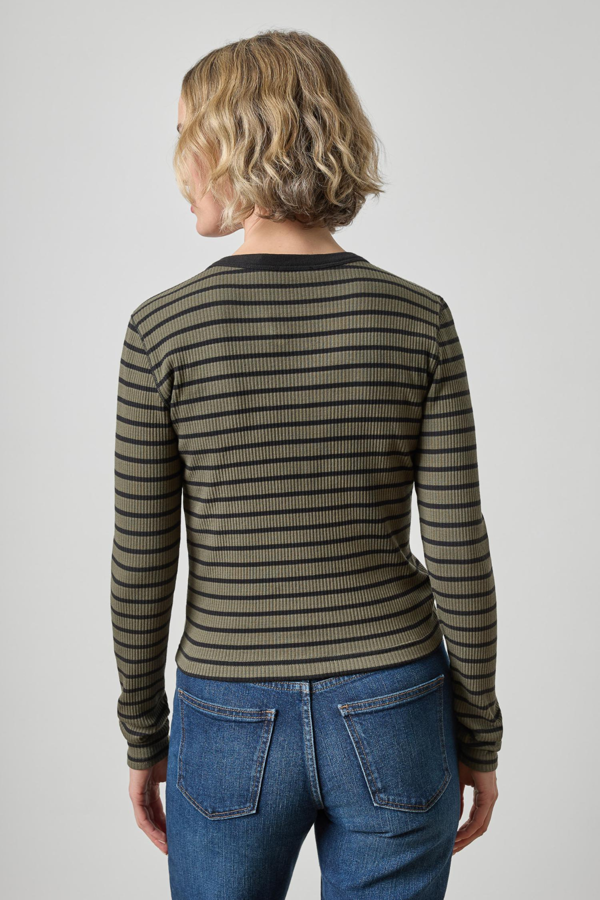 A woman viewed from the back wearing a long-sleeve ribbed knit top with horizontal olive green and black stripes, paired with blue jeans, against a light gray studio background.