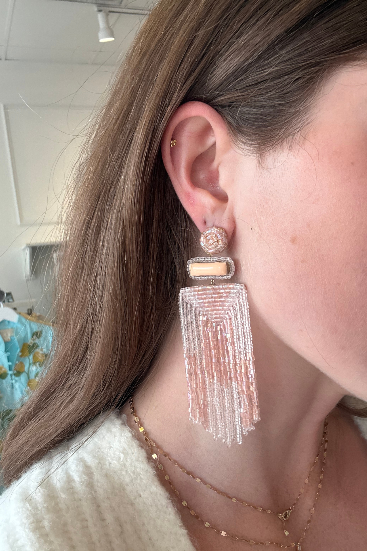 A close-up of a woman wearing a long, dangling statement earring. The earring features a gold-toned geometric setting with a small rectangular stone and a large, cascading fringe tassel made of clear and light pink beads.