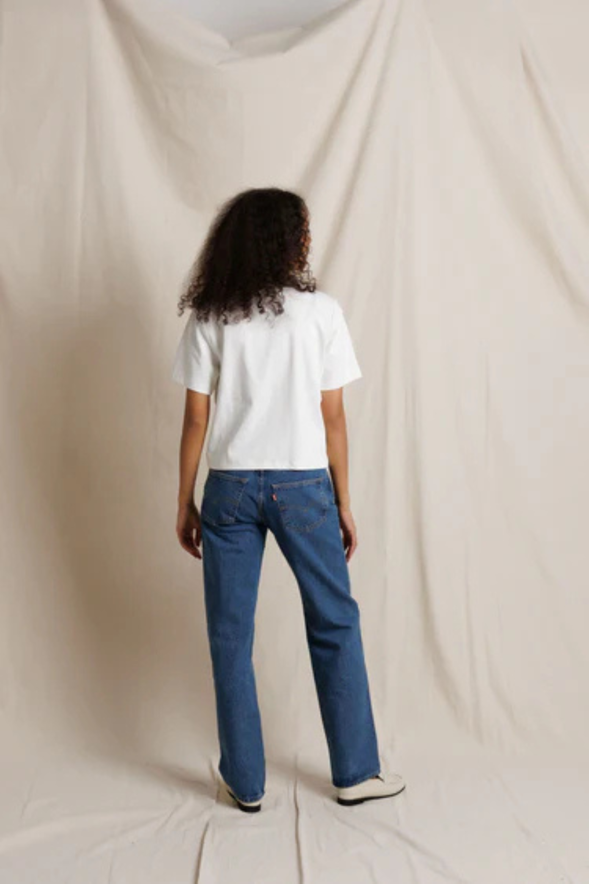 Person wearing a white t-shirt and blue jeans standing against a beige backdrop