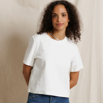 A woman with curly dark hair wearing a plain white short-sleeve crop top and blue jeans, standing against a draped beige fabric backdrop.