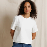 A woman with curly dark hair wearing a plain white short-sleeve crop top and blue jeans, standing against a draped beige fabric backdrop.