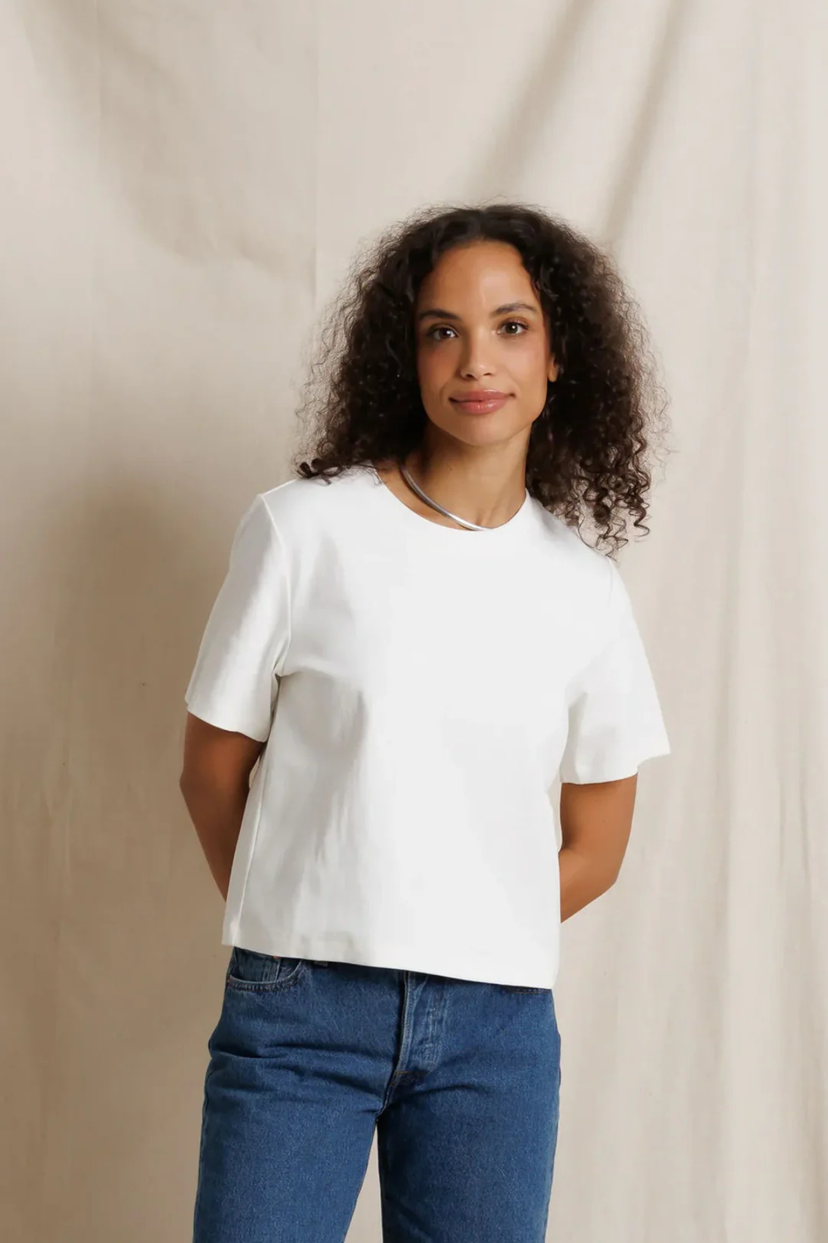 A woman with curly dark hair wearing a plain white short-sleeve crop top and blue jeans, standing against a draped beige fabric backdrop.